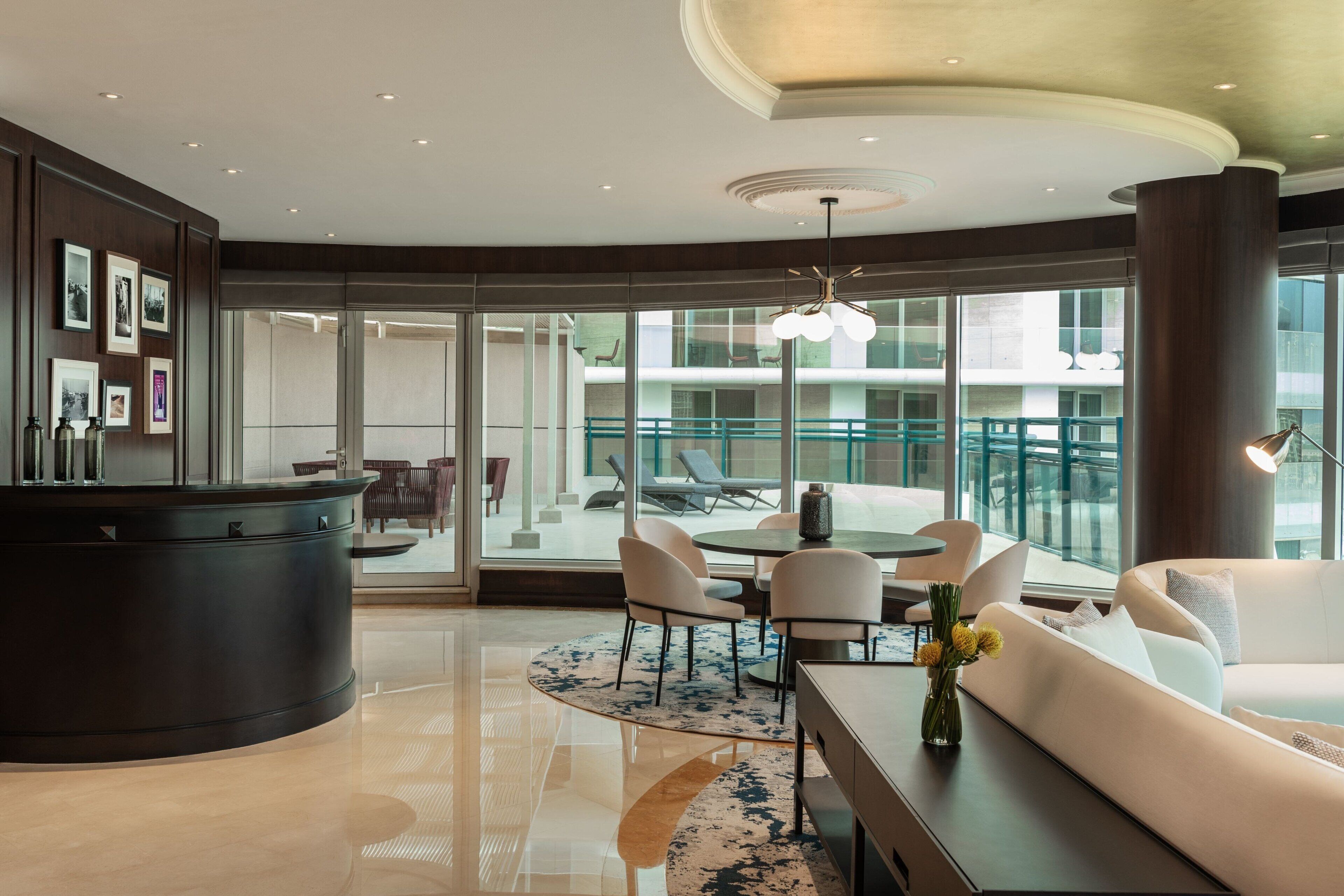 A modern hotel lobby with a round dining table, white sofas, and a large window offering a view of the outdoor seating area.
