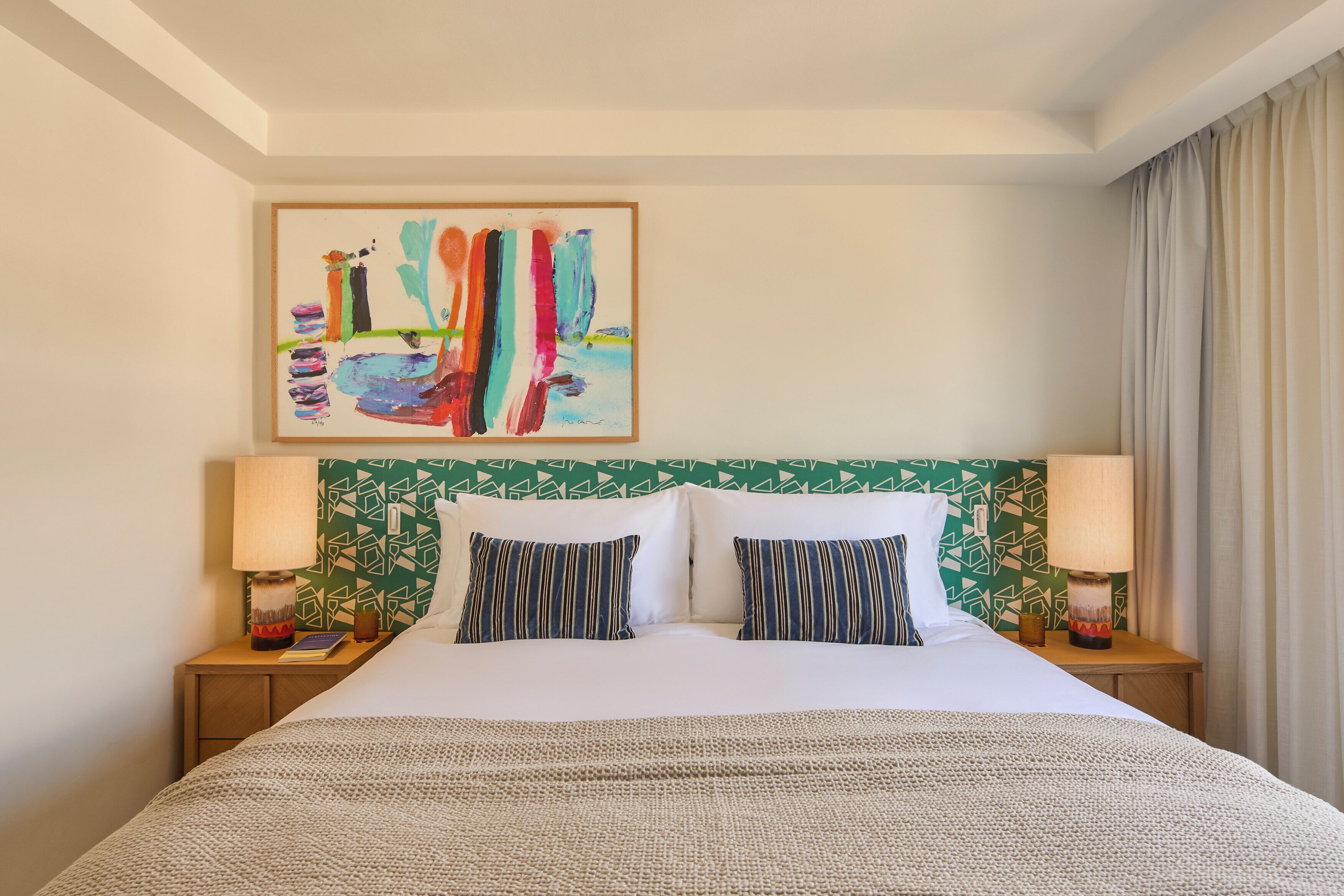 A hotel room with a bed, two pillows, a headboard with a geometric pattern, a painting on the wall, and two bedside lamps.