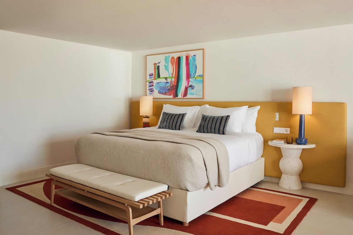 A neatly arranged hotel room with a bed, bedside tables, a bench, and a painting on the wall.