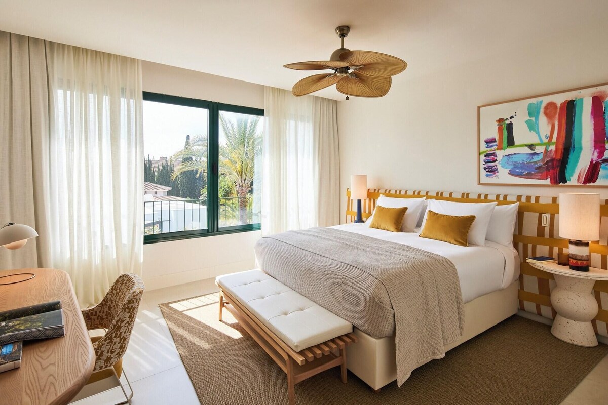 A hotel room with a bed, a bench, a desk, a chair, a ceiling fan, a window with curtains, and a painting on the wall.