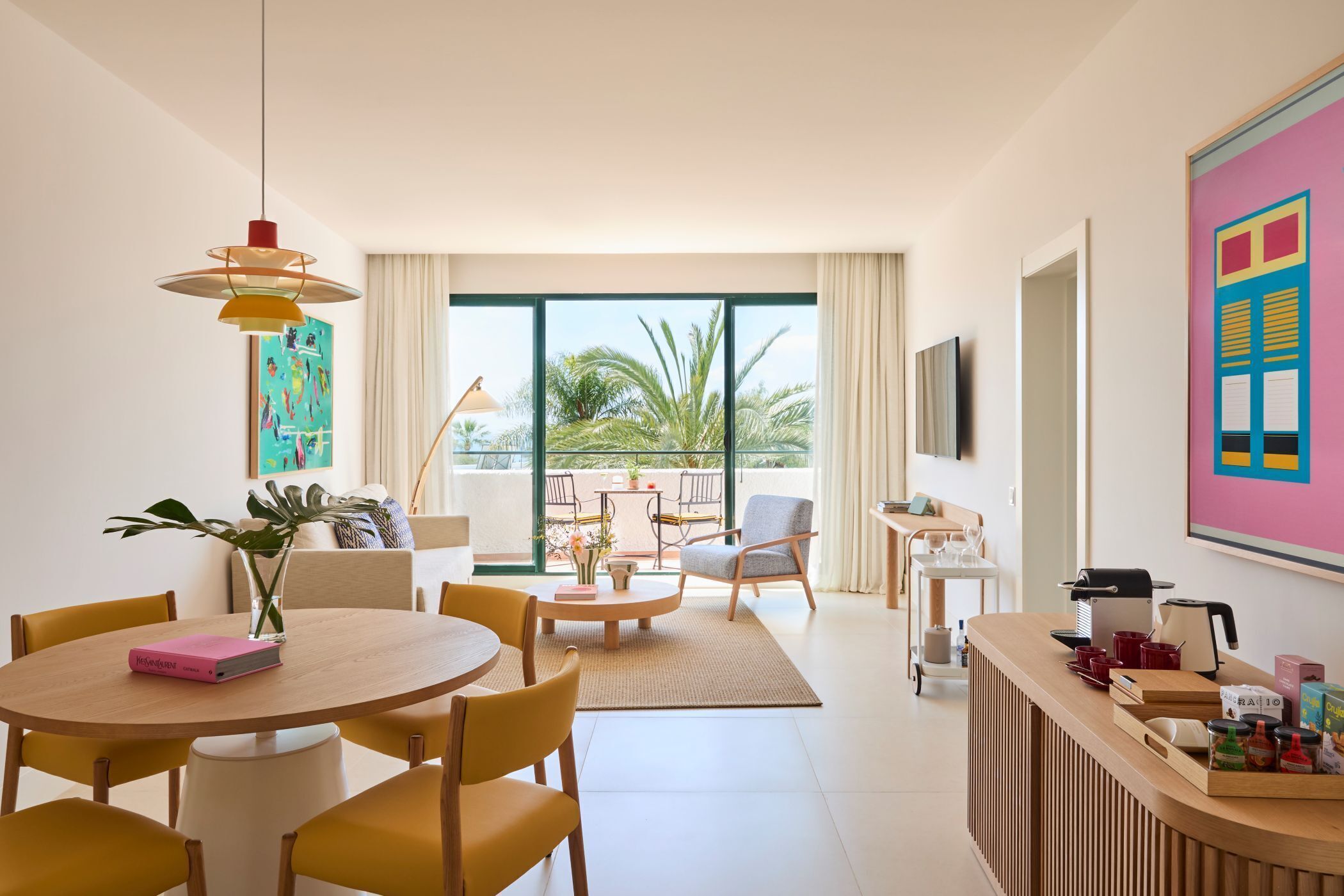 Een moderne hotelkamer met een zithoek, een bar, een groot raam met uitzicht op palmbomen en een wandgemonteerde flatscreen televisie.