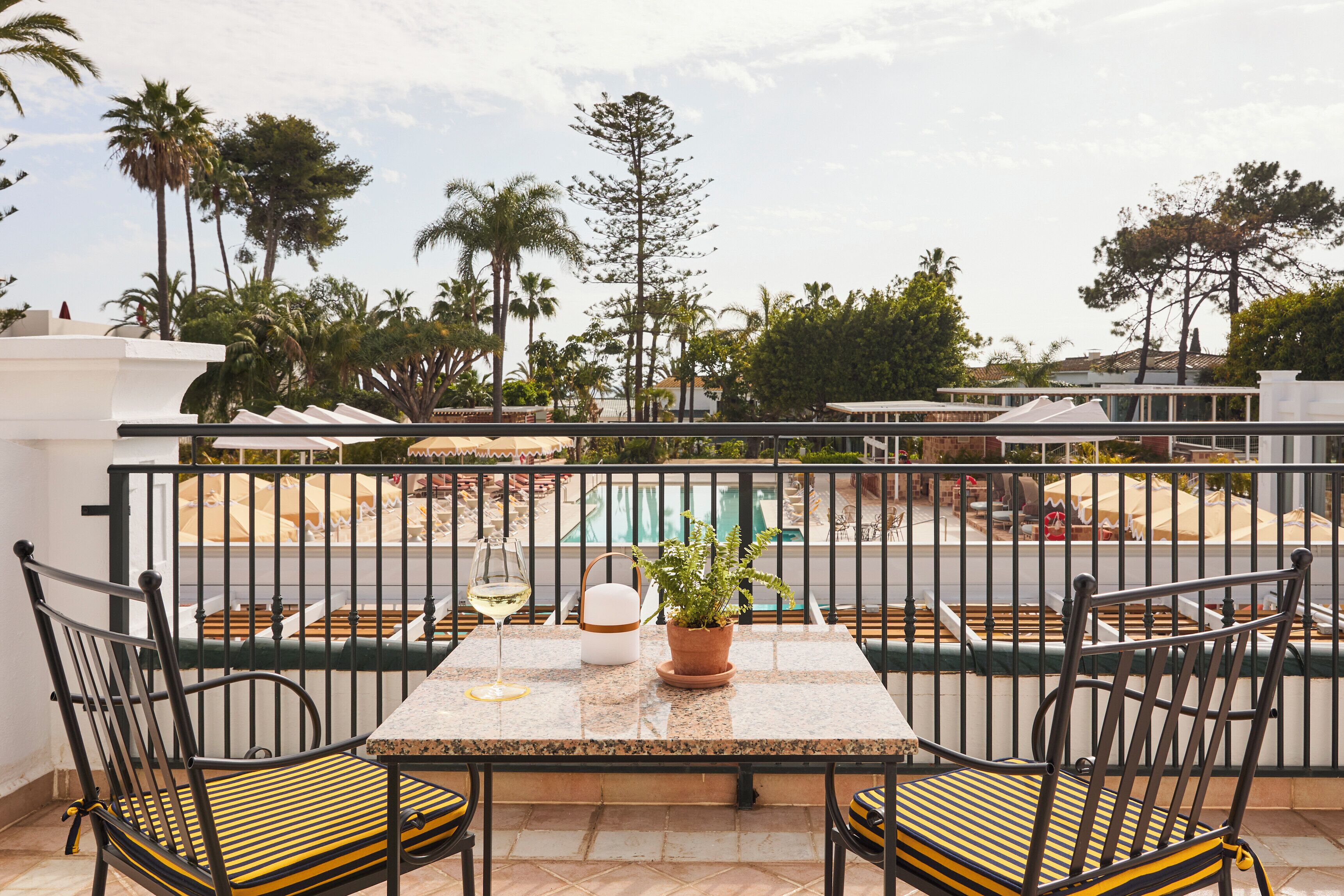 A balcony with a table, chairs, a potted plant, and a glass of wine.