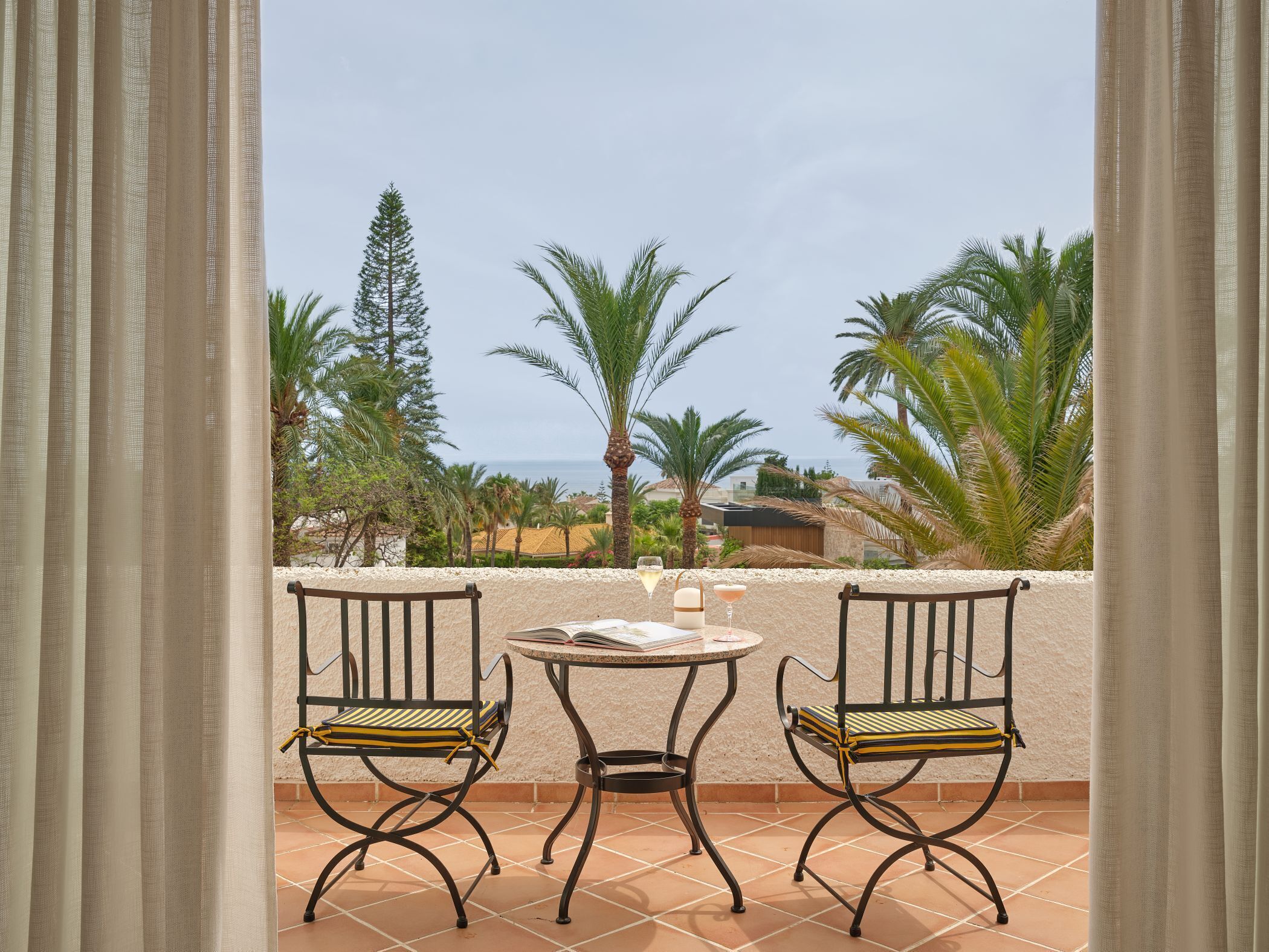 Un balcon avec deux chaises et une table, donnant sur un paysage côtier avec des palmiers et une vue sur la mer.