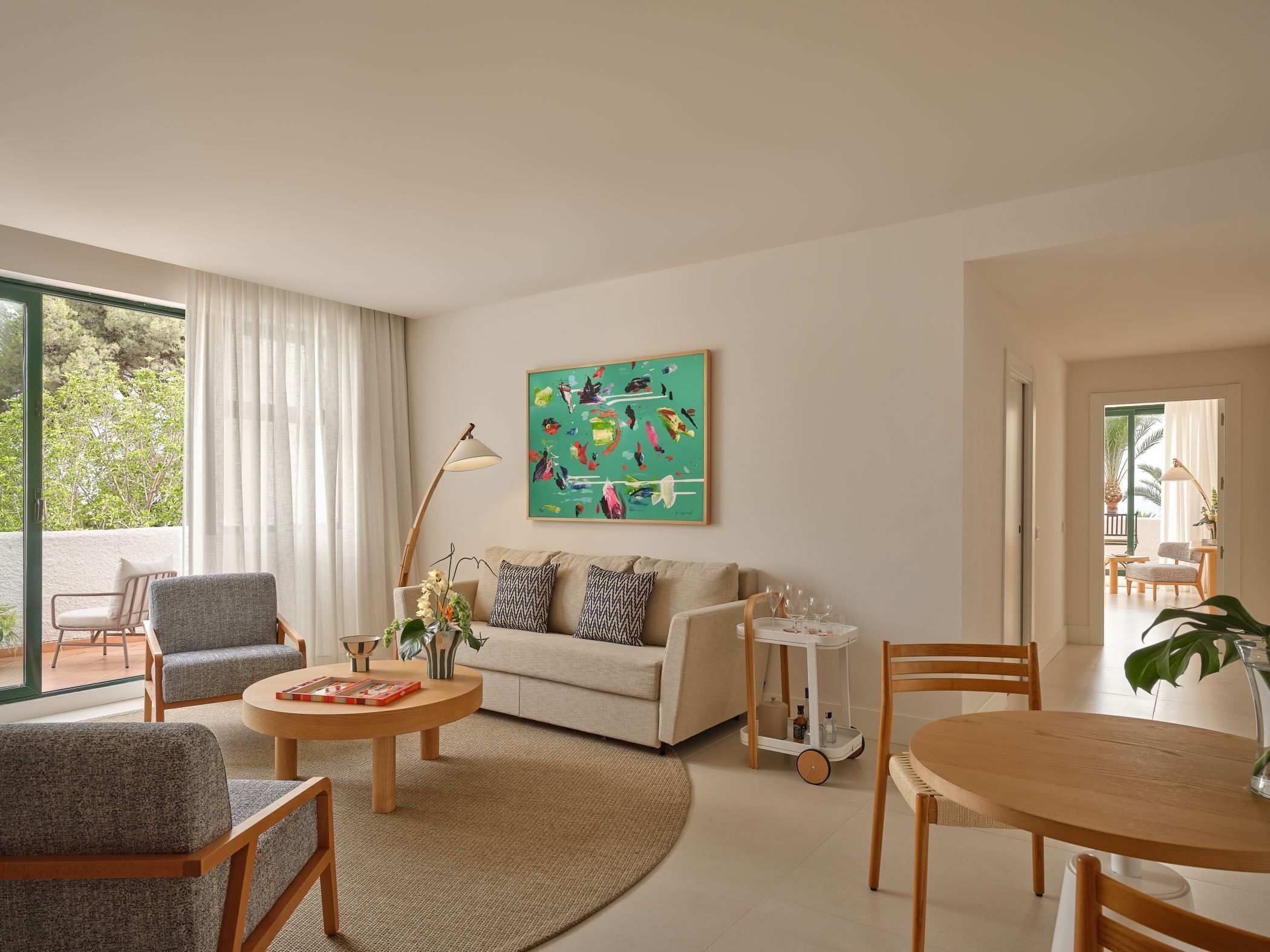 Un salon moderne comprenant un canapé, deux fauteuils, une table basse et une table à manger. Un tableau orne le mur, une lampe sur pied est posée au sol, et une grande fenêtre offre une vue sur l’extérieur.