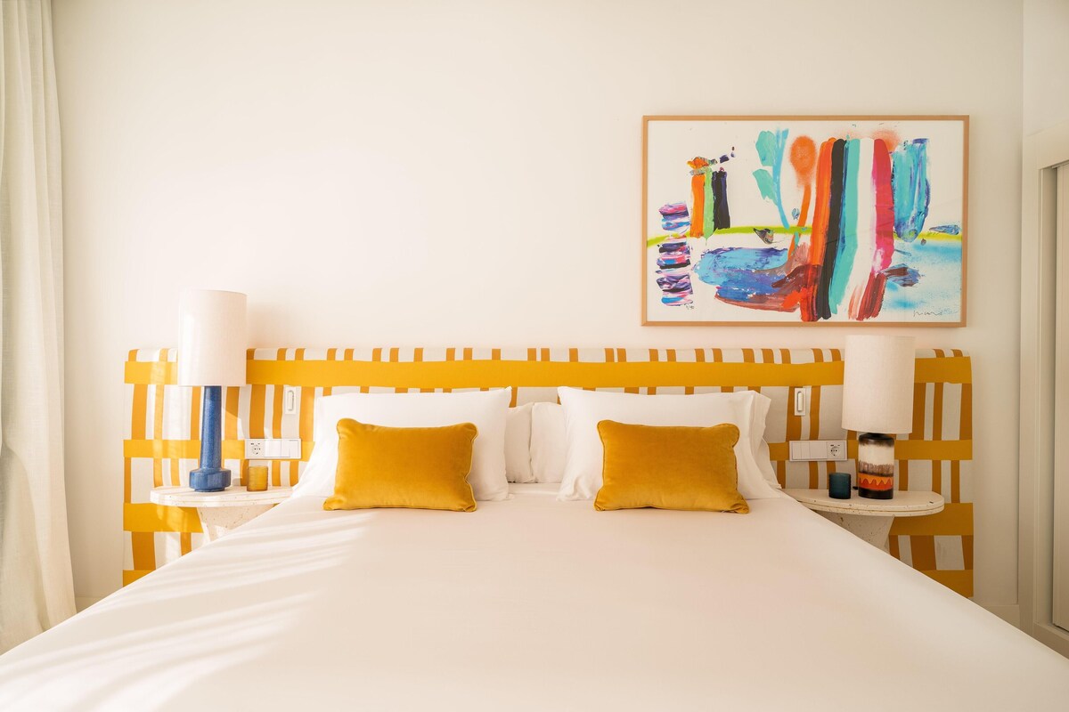 A hotel room with a bed, two bedside tables, a lamp, and a colorful abstract painting on the wall.