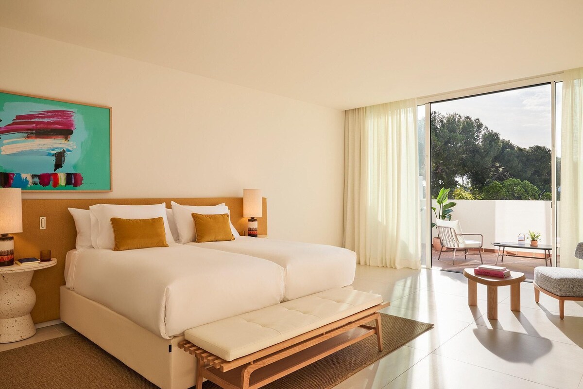 A hotel room with a large bed, a bedside table, a lamp, a painting, and a balcony with outdoor furniture.