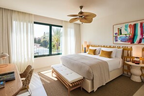 Habitación de hotel con una cama grande, un banco, un escritorio y vistas a palmeras.