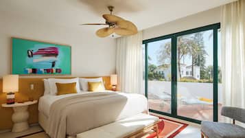 A hotel room with a bed, a chair, a ceiling fan, a window with a view of palm trees, and a painting on the wall.