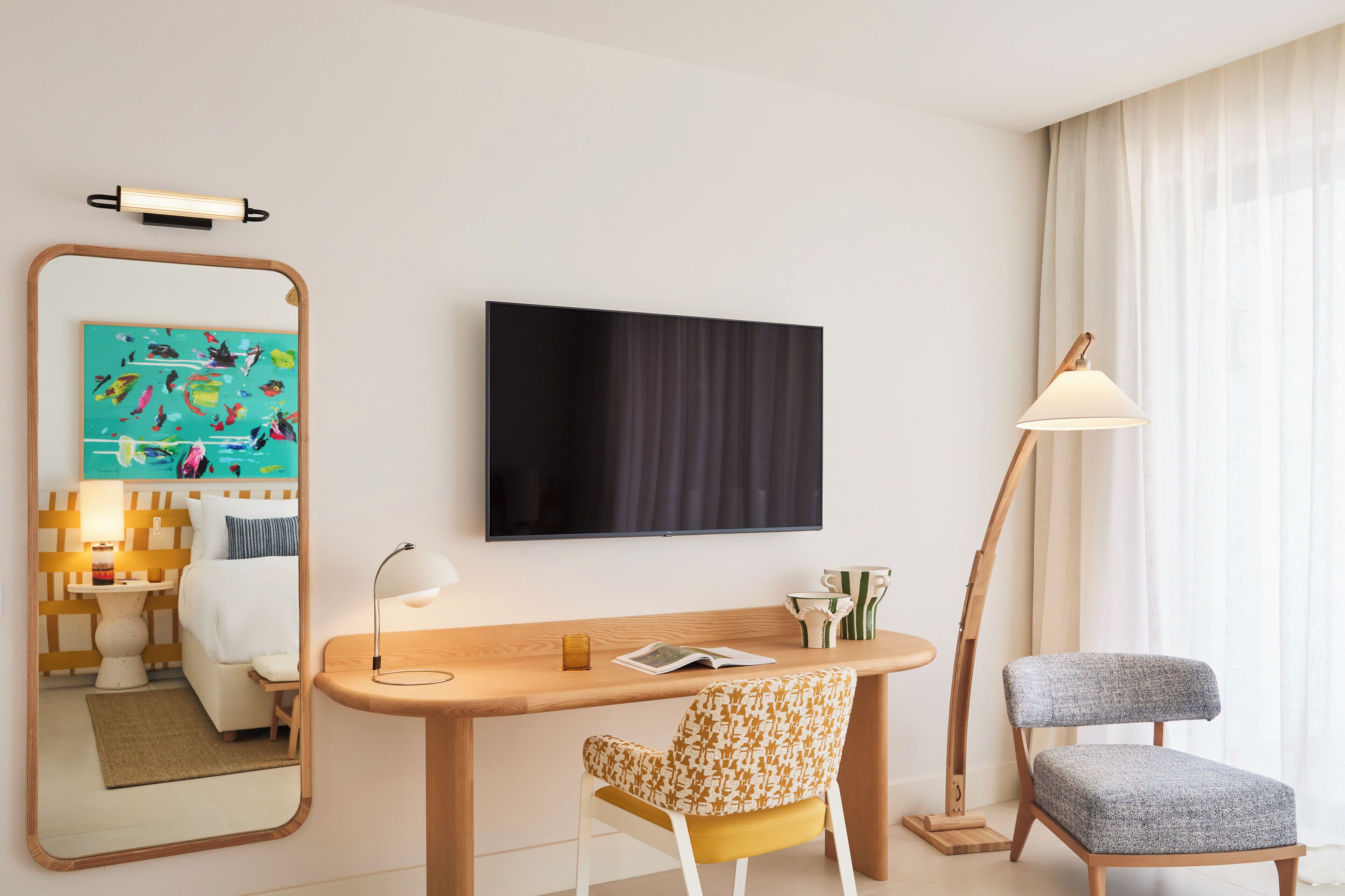 A hotel room with a wooden desk, a chair, a large mirror, a flat-screen TV, and a lamp.