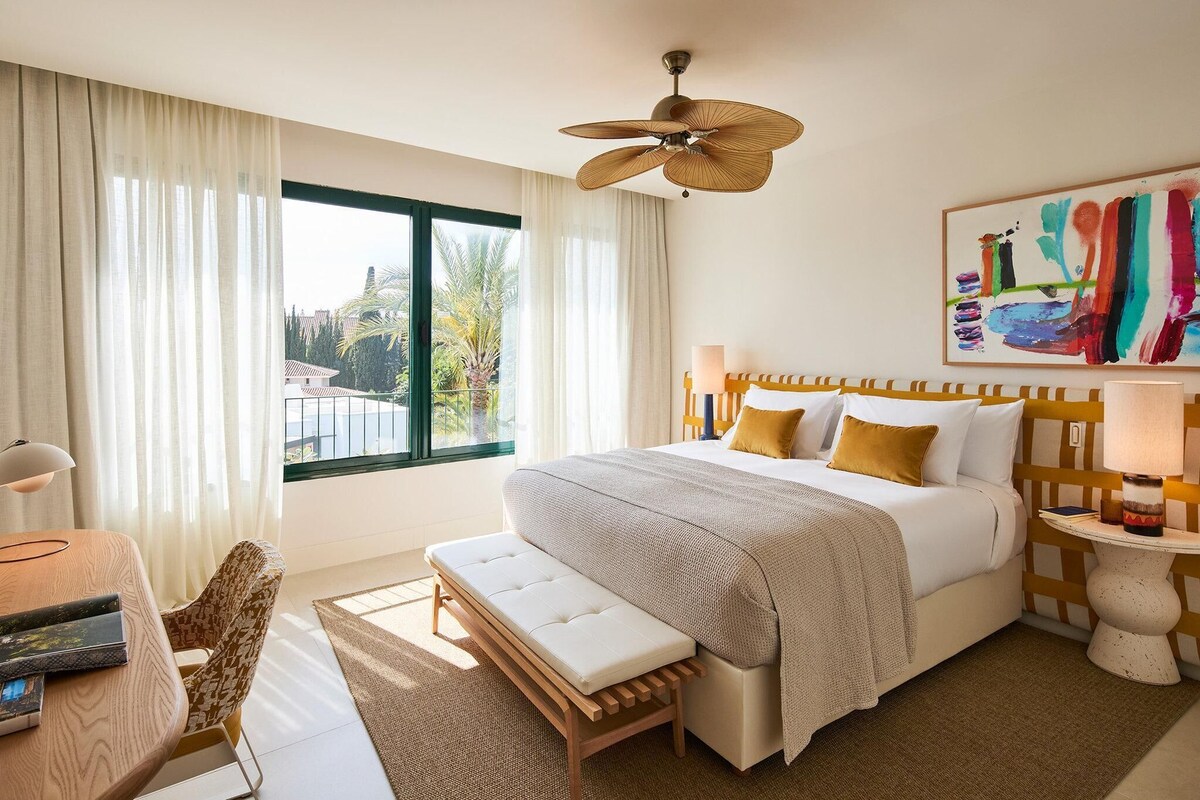 A hotel room with a bed, a bench, a desk, a chair, a ceiling fan, a window with curtains, and a painting on the wall.