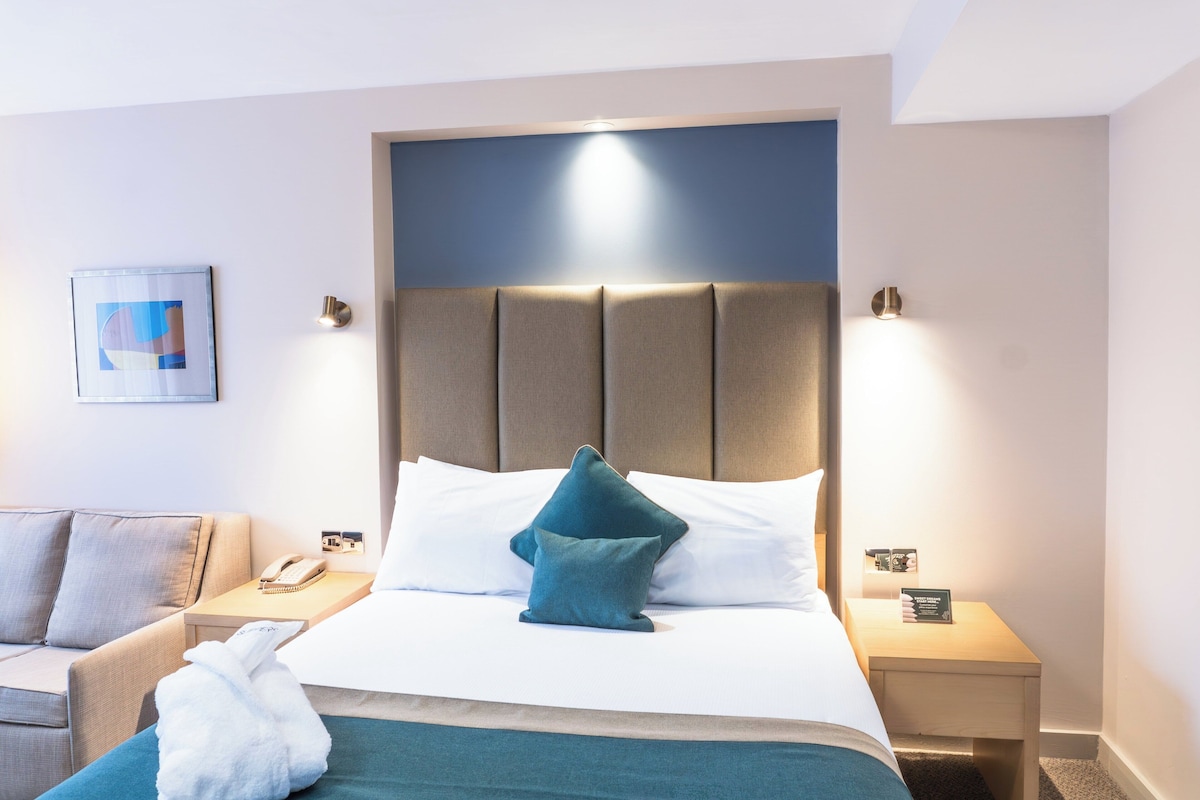 Ein Hotelzimmer mit Bett, Nachttisch, Lampe, einem fest montierten Telefon und einem gerahmten Bild.