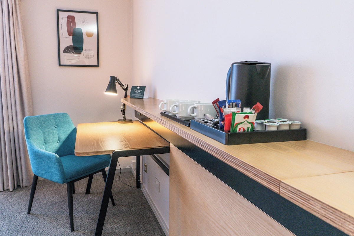 Habitación de hotel con escritorio de madera, una silla color turquesa, cafetera y un cuadro abstracto enmarcado en la pared.
