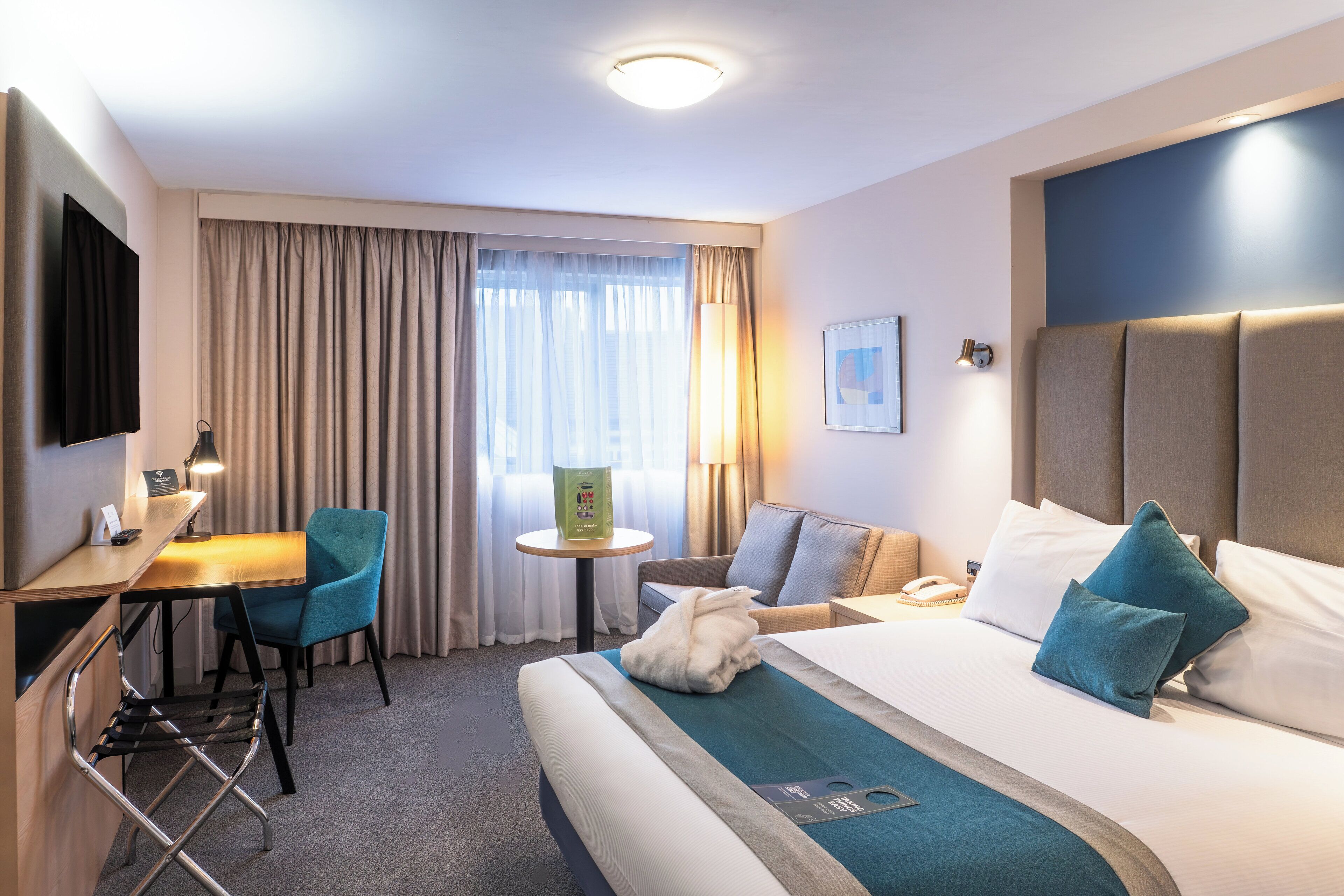 Habitación de hotel con una cama, un escritorio con una silla, una mesita y un televisor instalado en la pared.