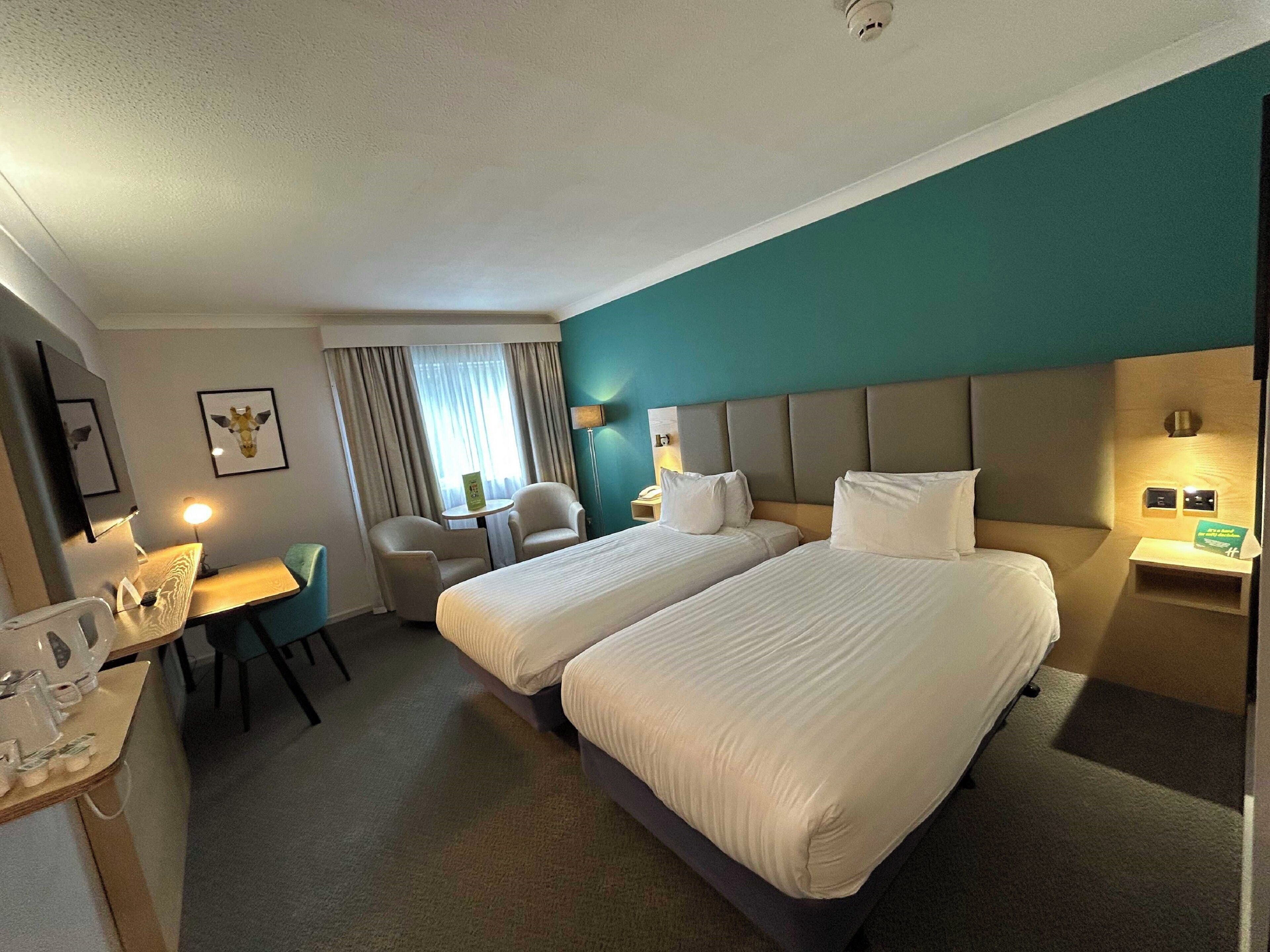 Een hotelkamer met twee bedden, een bureau, een stoel, een kleine tafel en een raam met gordijnen.