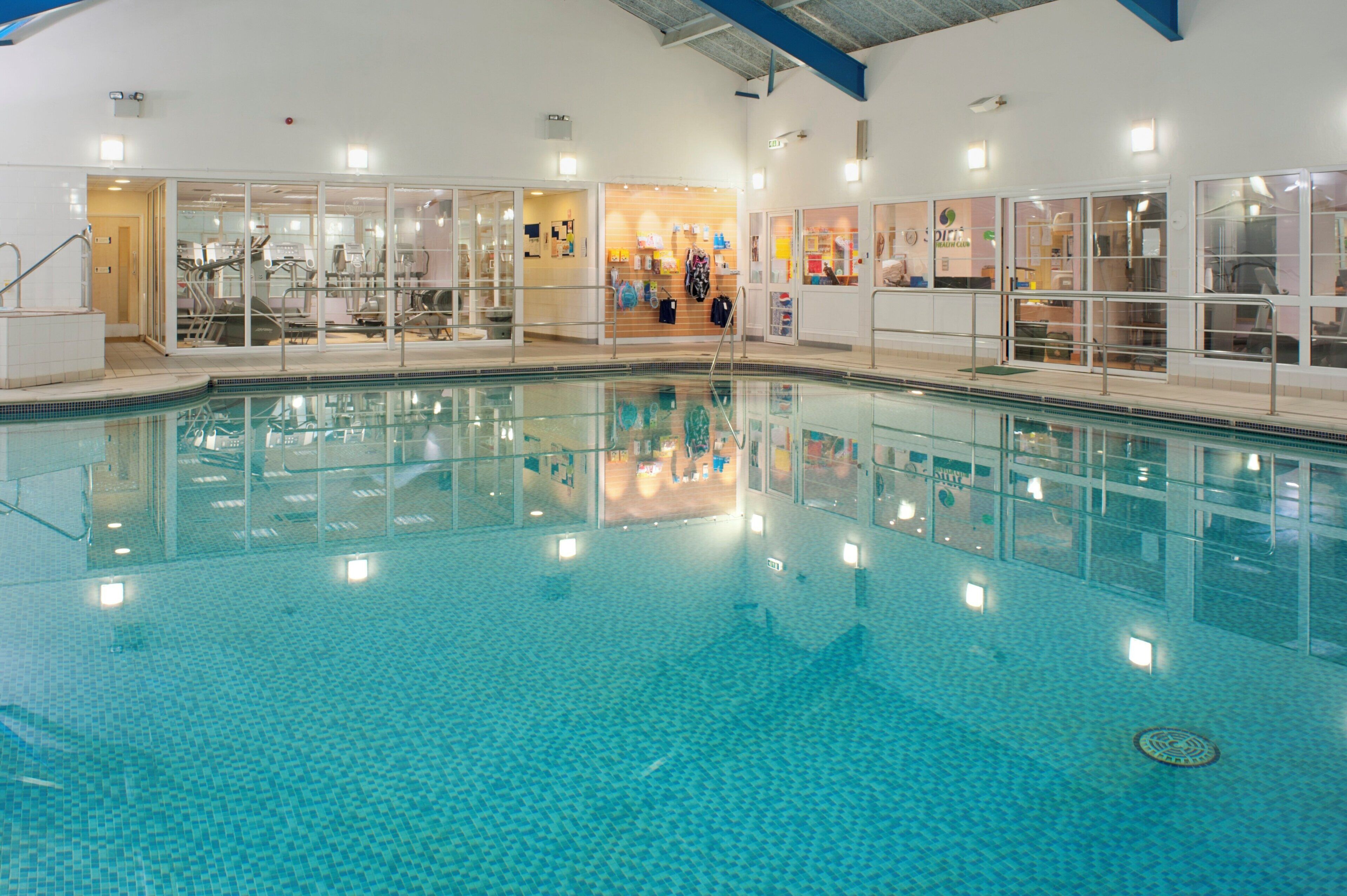 An indoor swimming pool with clear water, surrounded by glass walls, and various amenities visible through the glass.