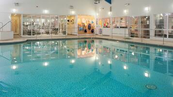 An indoor swimming pool with clear water, surrounded by glass walls, and various amenities visible through the glass.