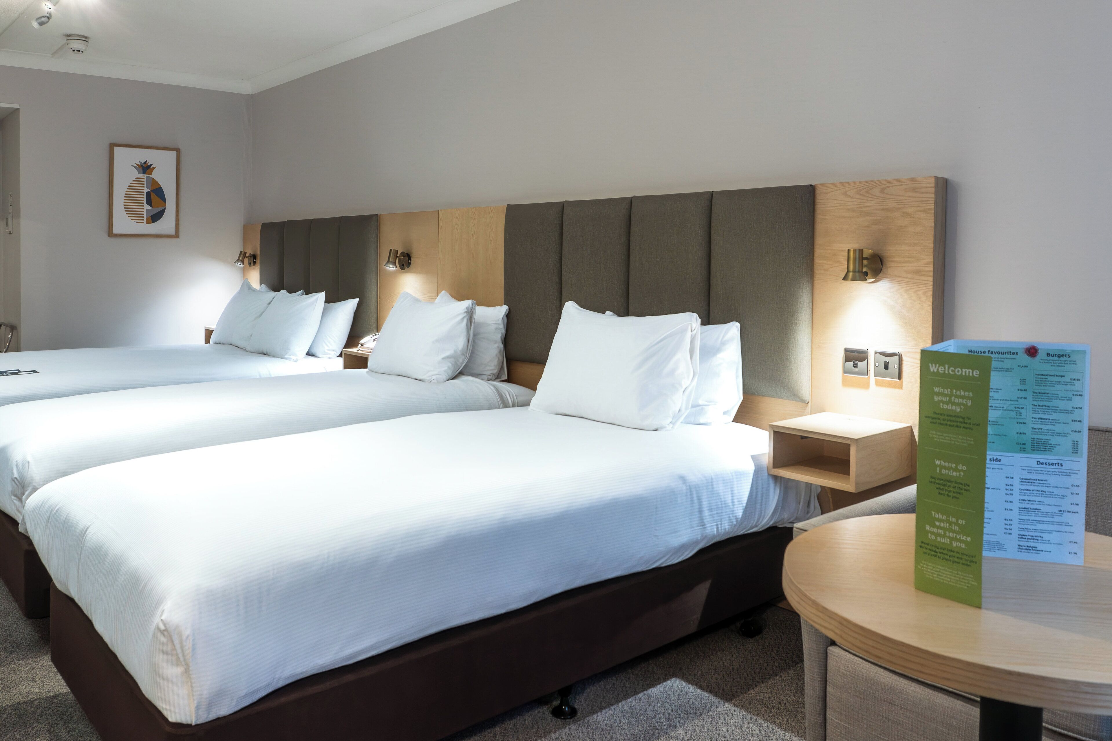 A hotel room with two beds, a wooden headboard, bedside tables, and a round table with a menu stand.