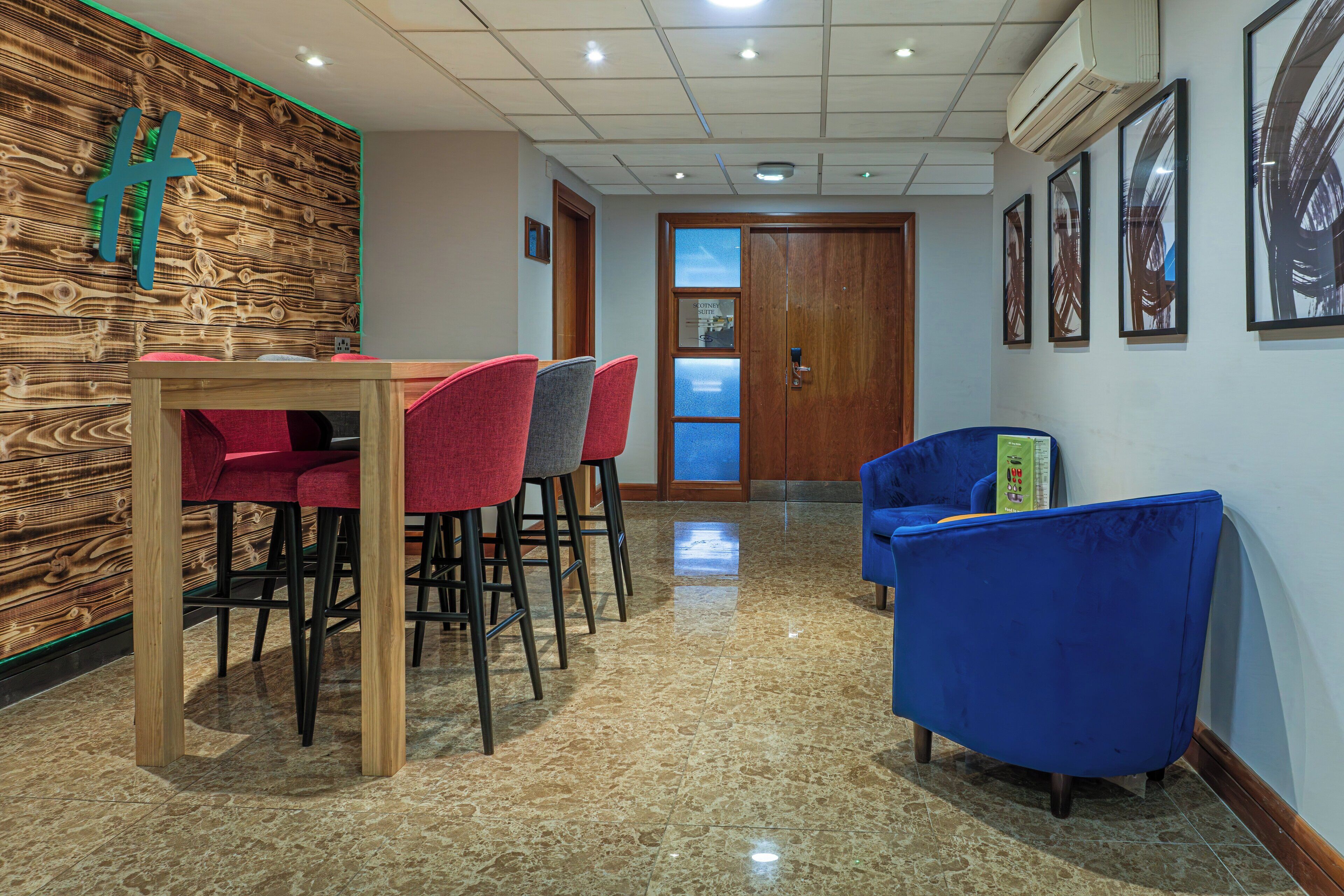Een receptiezone met een houten wand, een receptiebalie, rode en grijze stoelen, een blauwe fauteuil en ingelijste kunstwerken aan de muur.