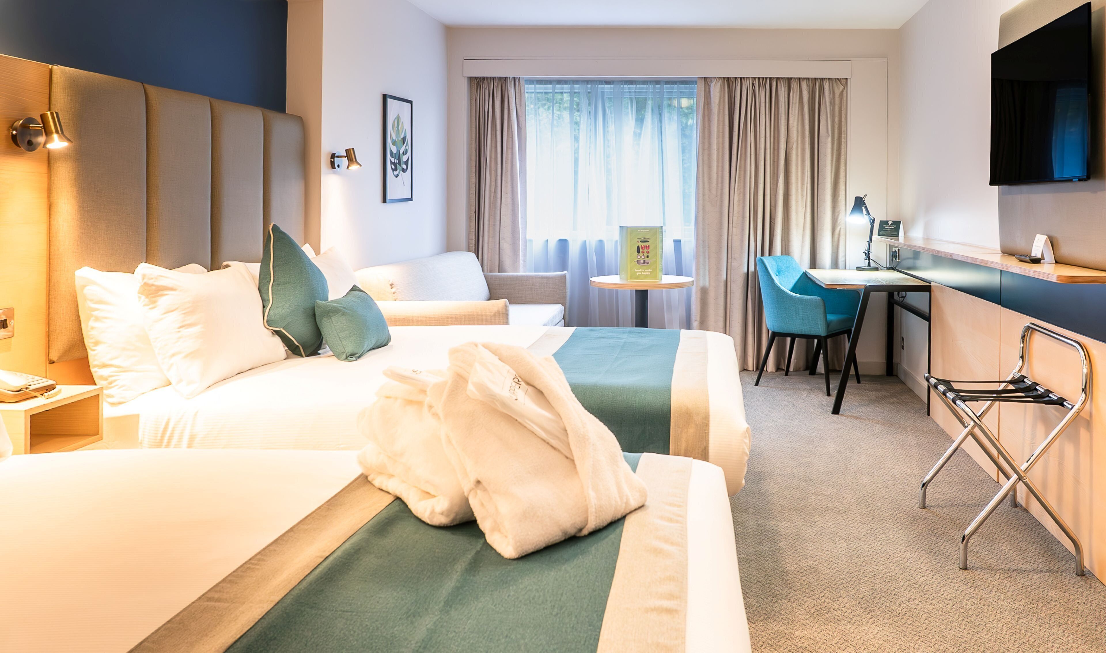 A hotel room with two beds, a desk, a chair, a TV, and a window with curtains.