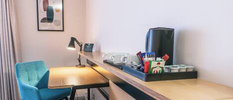 A hotel room with a wooden desk, a teal chair, a coffee maker, and a framed abstract art piece on the wall.