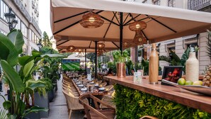 Un espace extérieur aménagé pour les repas, avec des chaises et des tables en osier, entouré de plantes en pot et de parasols.