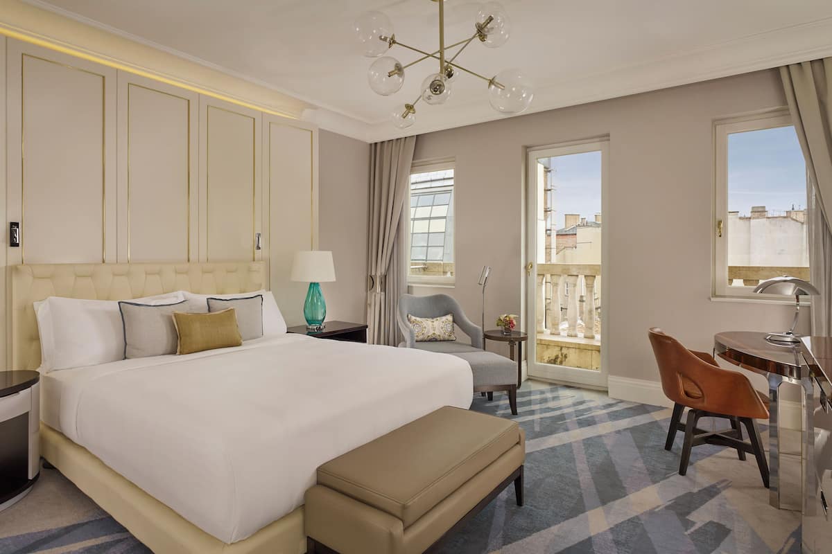 A hotel room with a large bed, two bedside tables with lamps, a chair, a small table, and a balcony with a view.