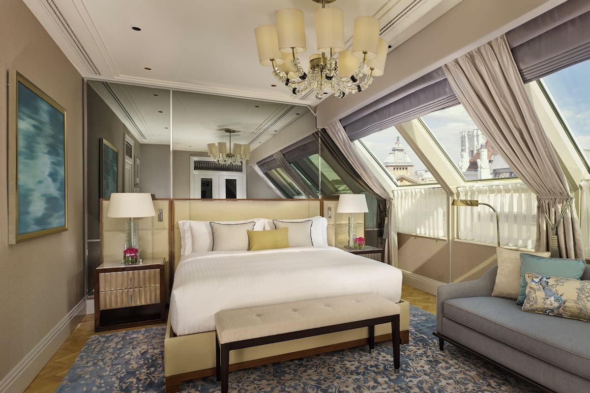 A spacious hotel room with a large bed, a sofa, a bench, and a view of the city through a skylight.