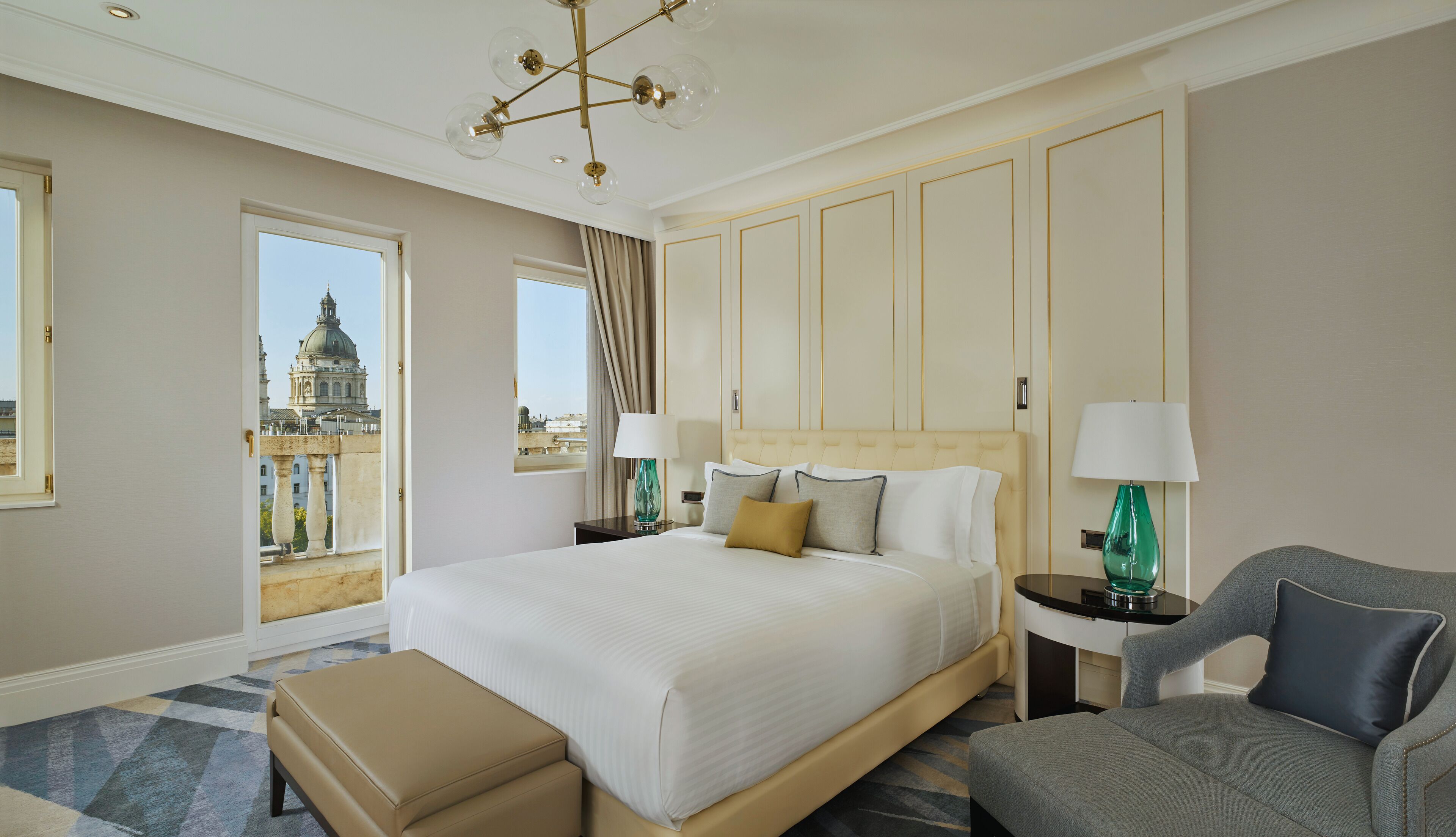 A hotel room with a large bed, two bedside tables with lamps, a chair, a bench, and a view of a historic building through a door.