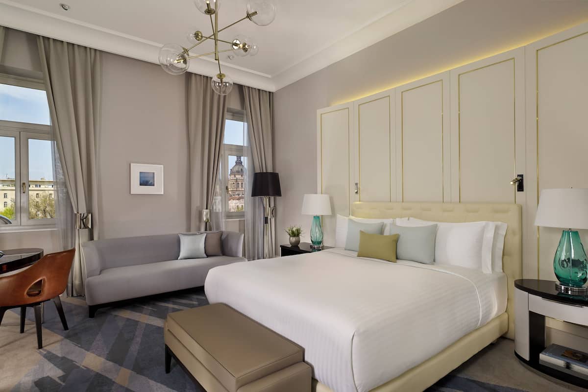 A hotel room with a large bed, a sofa, a desk, and a chair. There are two lamps on the bedside tables and a chandelier above the bed.