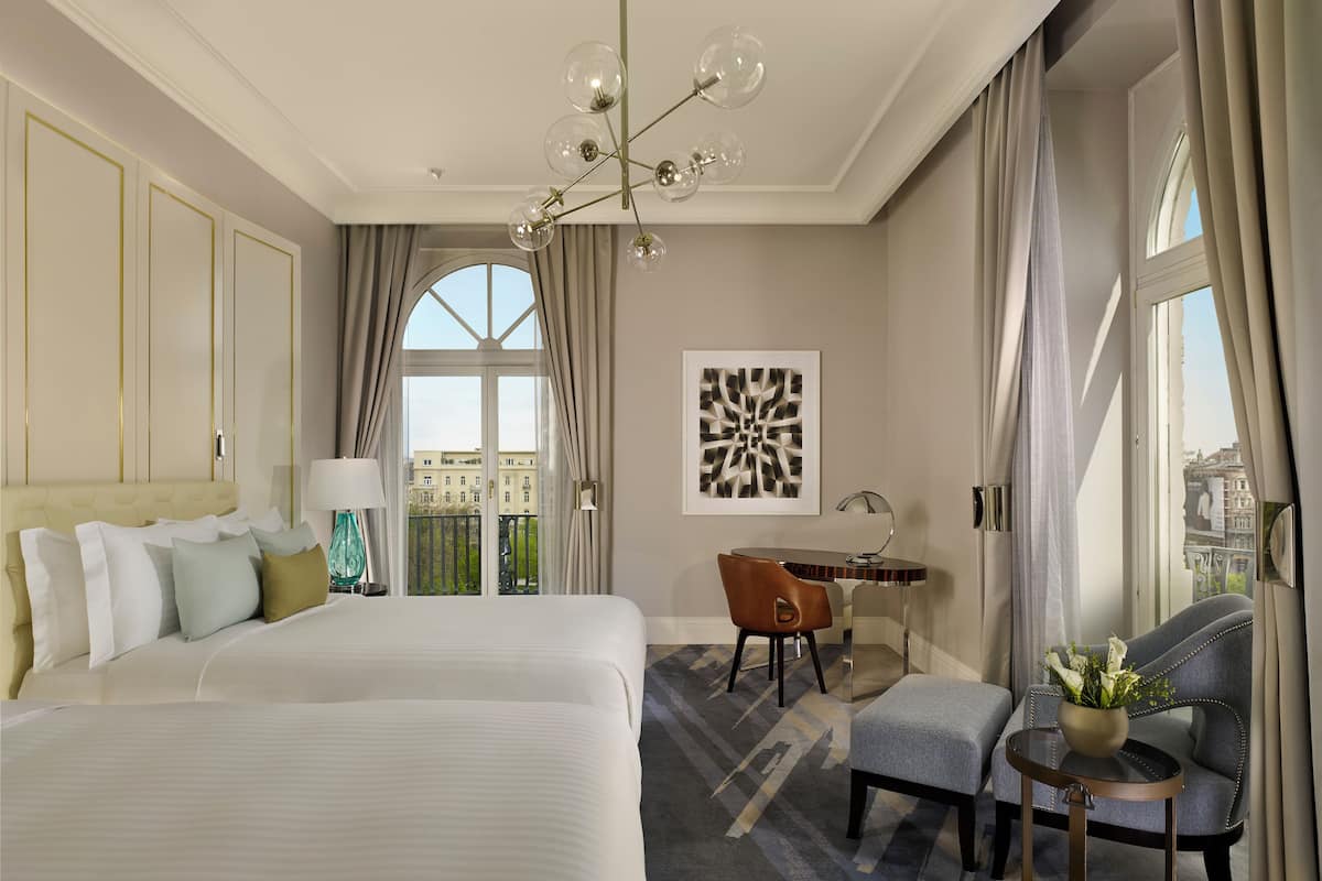 A hotel room with a large bed, a desk, a chair, a small table with a vase, and a view of the city through large windows.