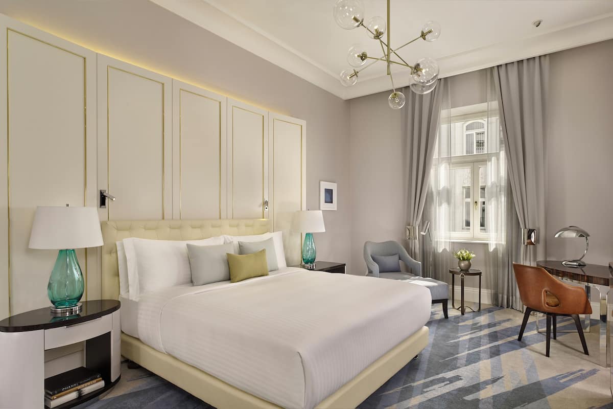 A hotel room with a large bed, bedside tables, a desk, a chair, and a chandelier.