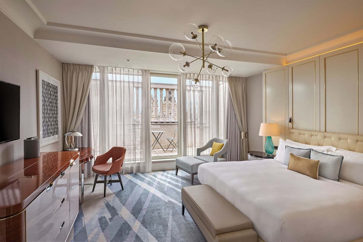 A hotel room with a large bed, a desk with a chair, a TV, and a balcony with a view of buildings.