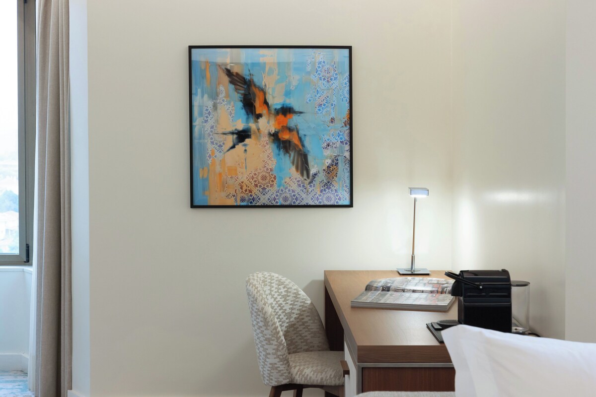 Habitación de hotel con una cama, un escritorio con lámpara, una silla y un cuadro abstracto en la pared.