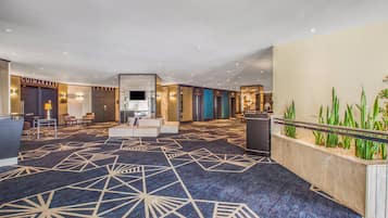 A hotel lobby with a geometric patterned carpet, seating areas, a reception desk, and a plant display.