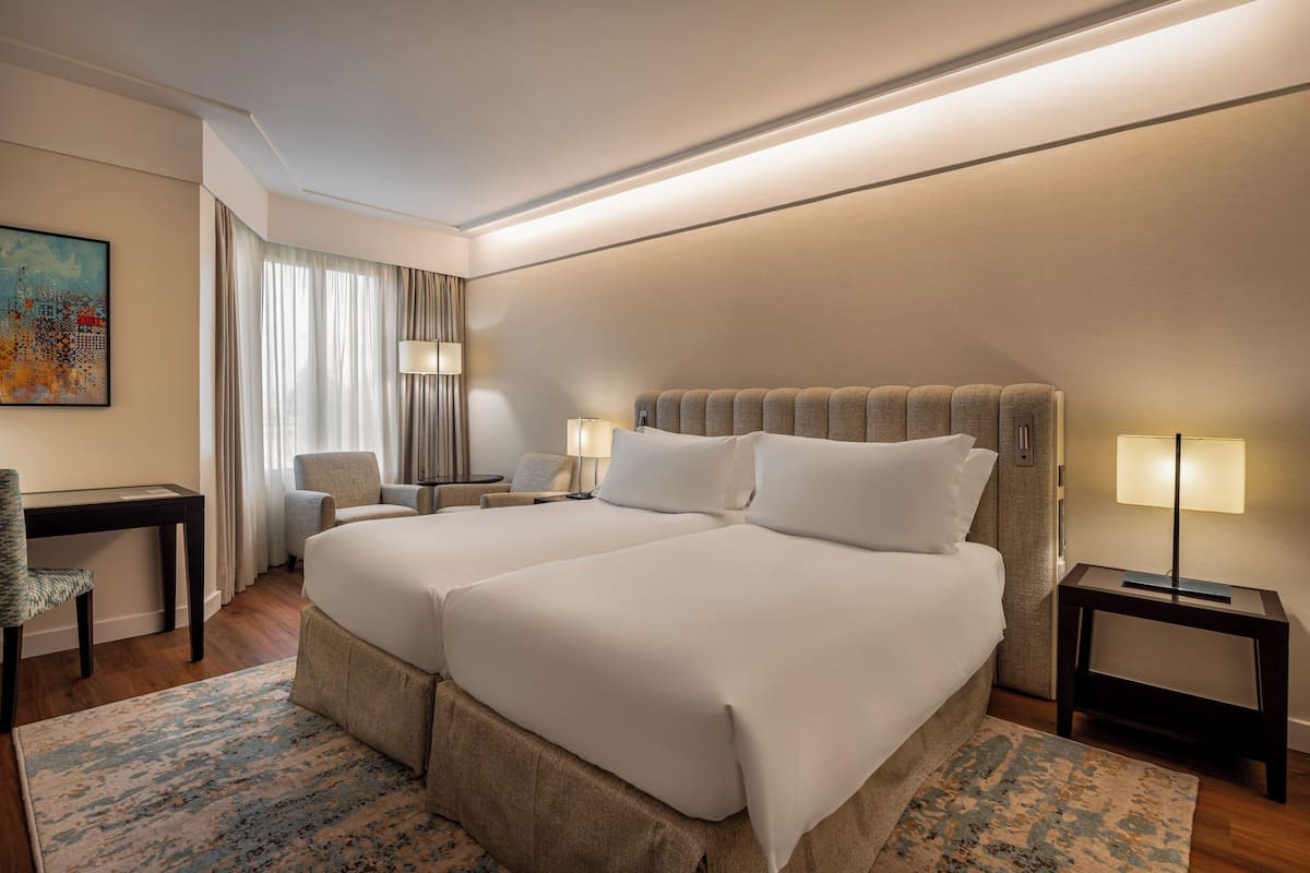 Habitación de hotel con dos camas, un escritorio, una silla, una lámpara y un cuadro en la pared.
