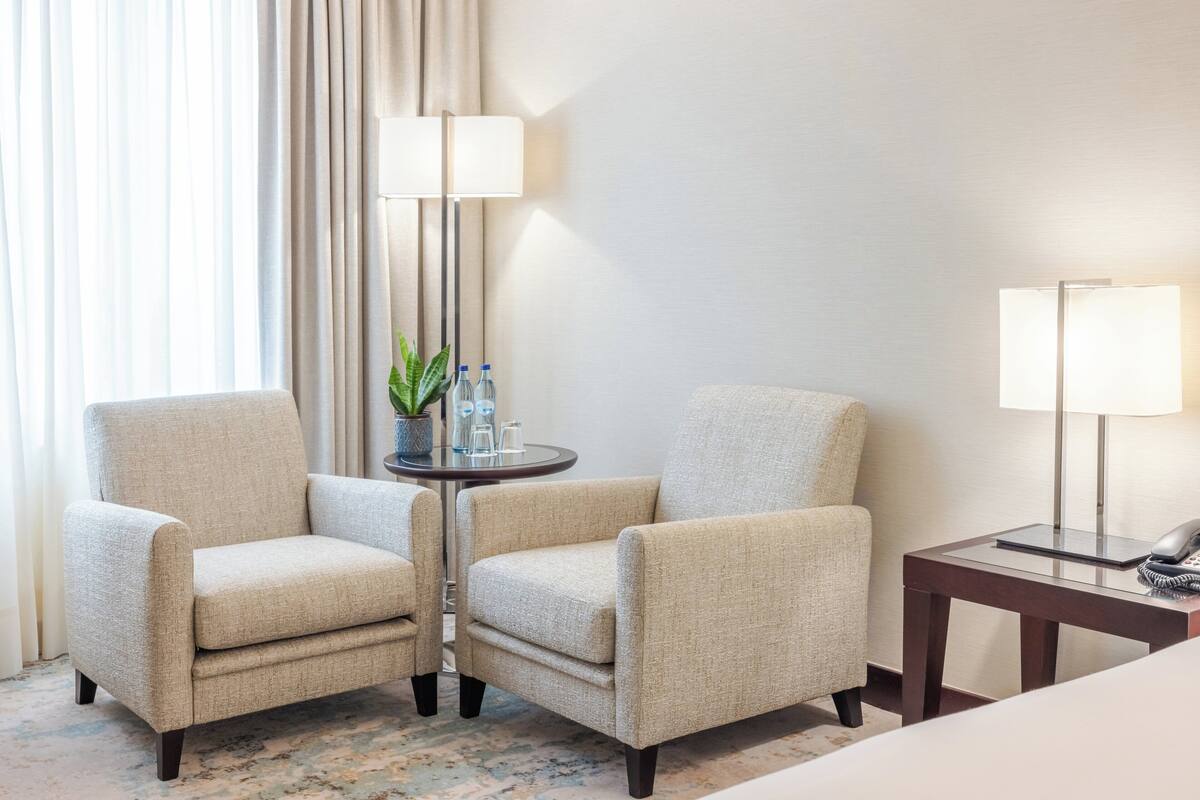 Habitación de hotel con dos sillones, una mesita con una planta y botellas de agua, una lámpara y una mesita de noche con un teléfono.