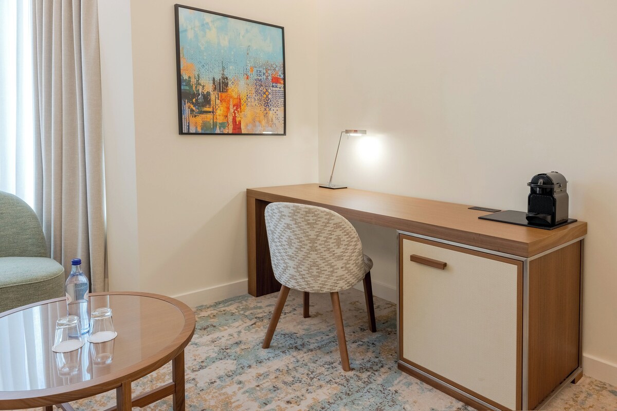 Una habitación de hotel con un escritorio de madera, una silla, una mesita de vidrio con botellas de agua, una lámpara y un cuadro en la pared.