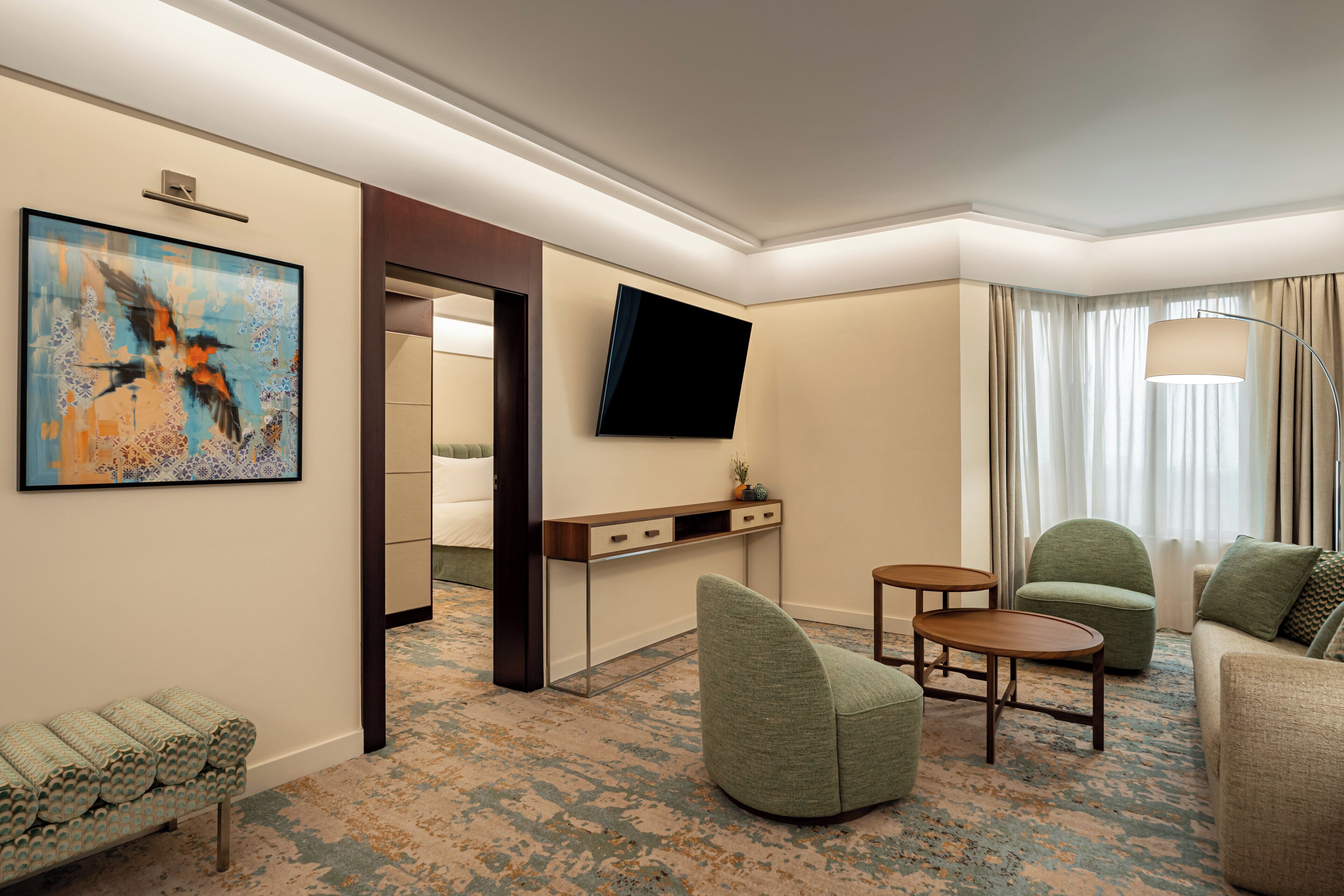 A hotel room with a flat-screen TV, a painting, a sofa, a chair, a small table, and a bed.