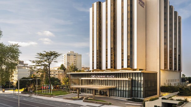 Un hotel moderno con un diseño vertical llamativo, un acceso cubierto y una zona ajardinada con árboles.