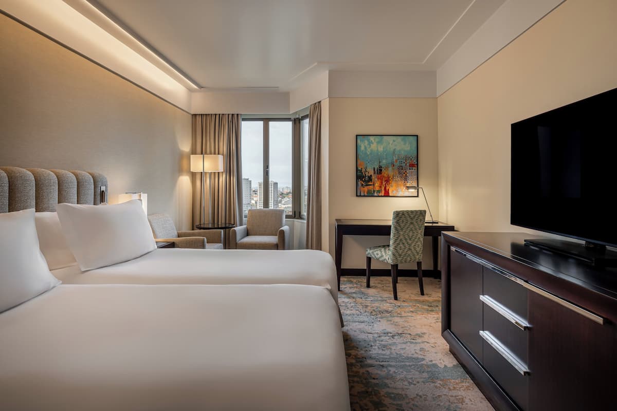 Habitación de hotel con dos camas, un escritorio, un televisor y vista a la ciudad.