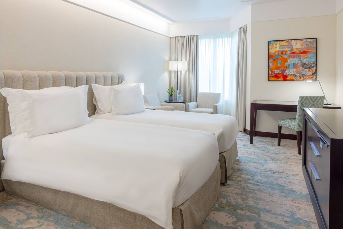 A hotel room with two beds, a desk, a chair, a lamp, and a painting on the wall.