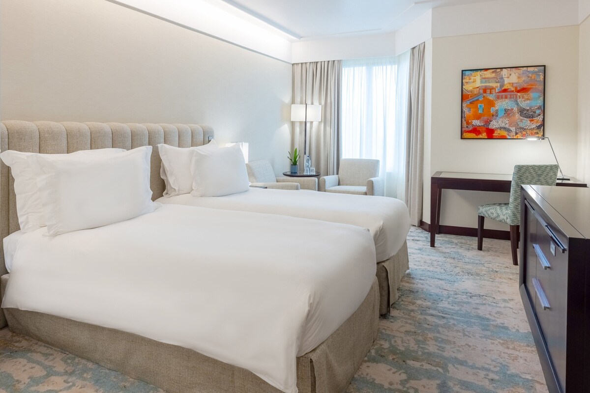 Habitación de hotel con dos camas, un escritorio, una silla, una lámpara y un cuadro en la pared.