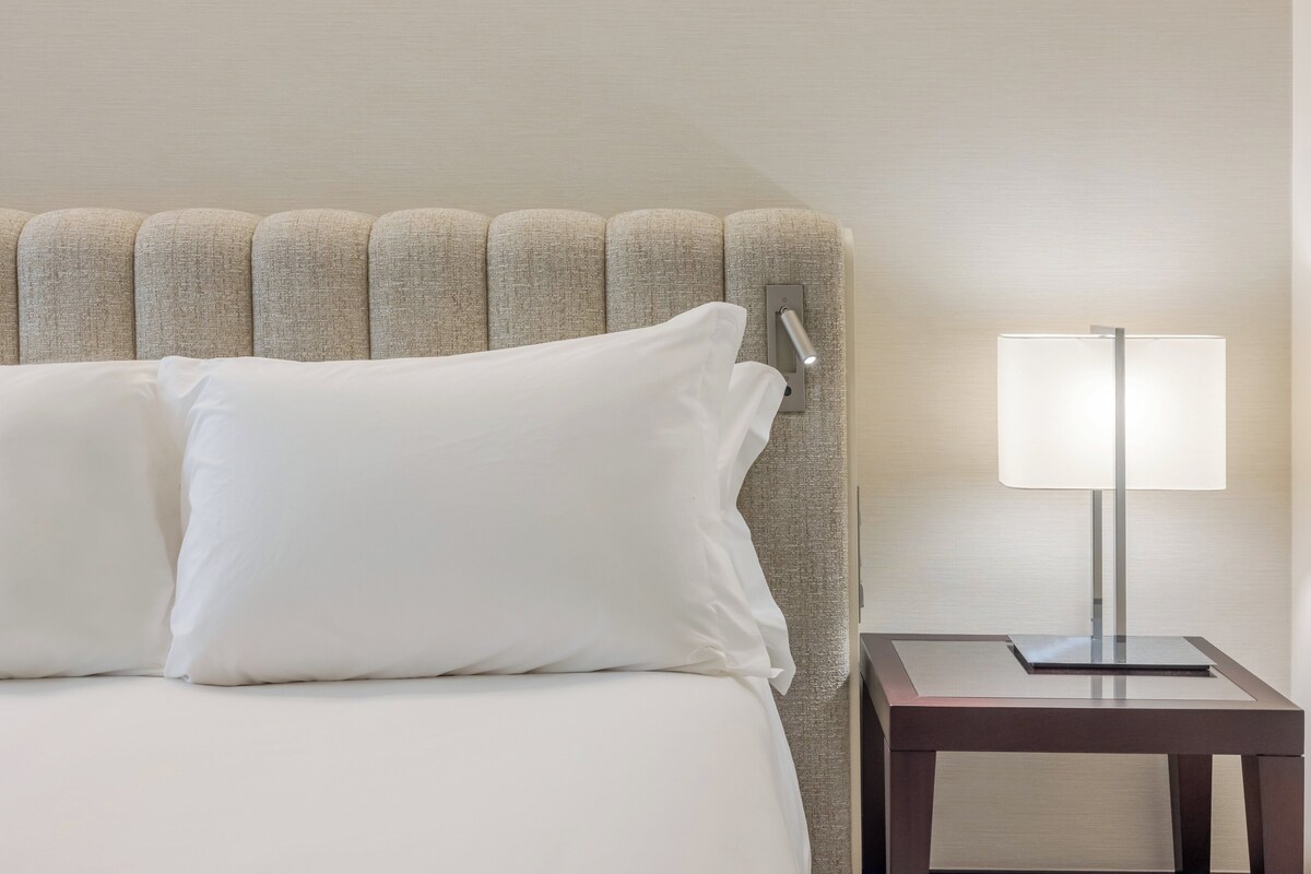 Una cama bien hecha con ropa blanca y una mesita de noche moderna con lámpara.