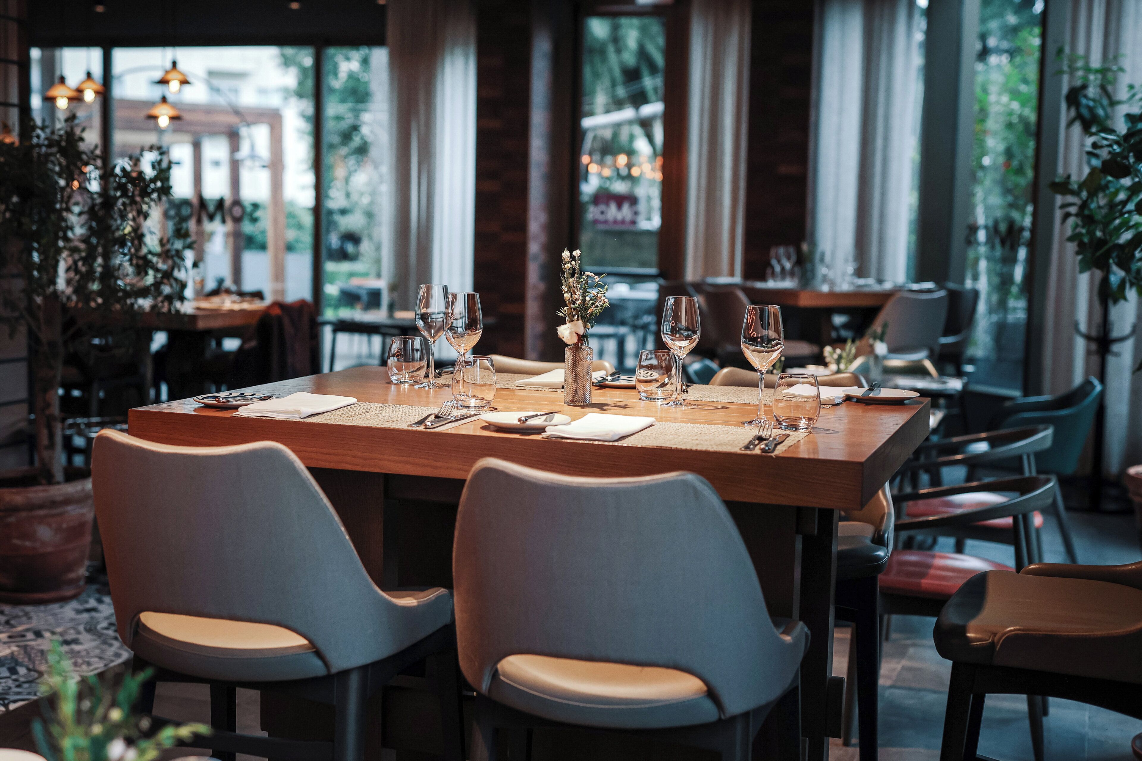 Une salle de restaurant avec une table en bois dressée pour le repas, plusieurs chaises et une vue dégagée sur l’extérieur grâce à de grandes fenêtres.