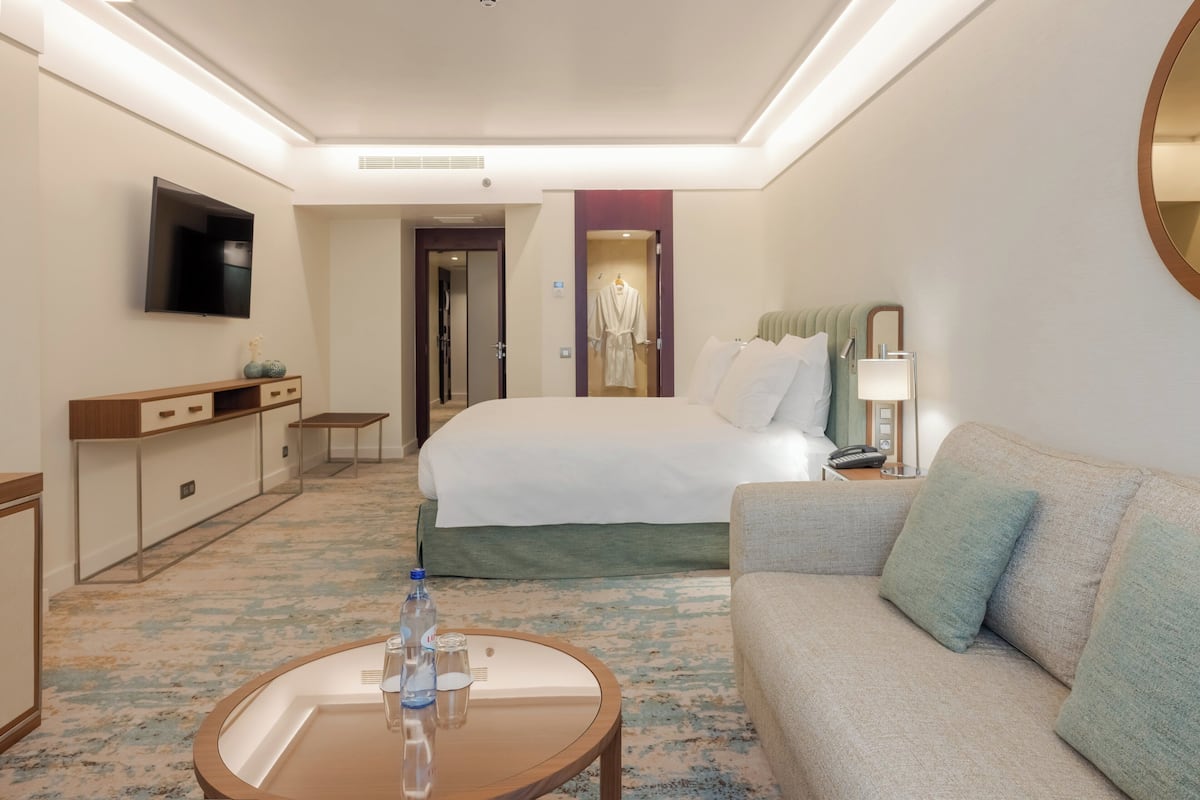 Habitación de hotel con cama, sofá, televisión, escritorio y baño visible a través de una puerta abierta.