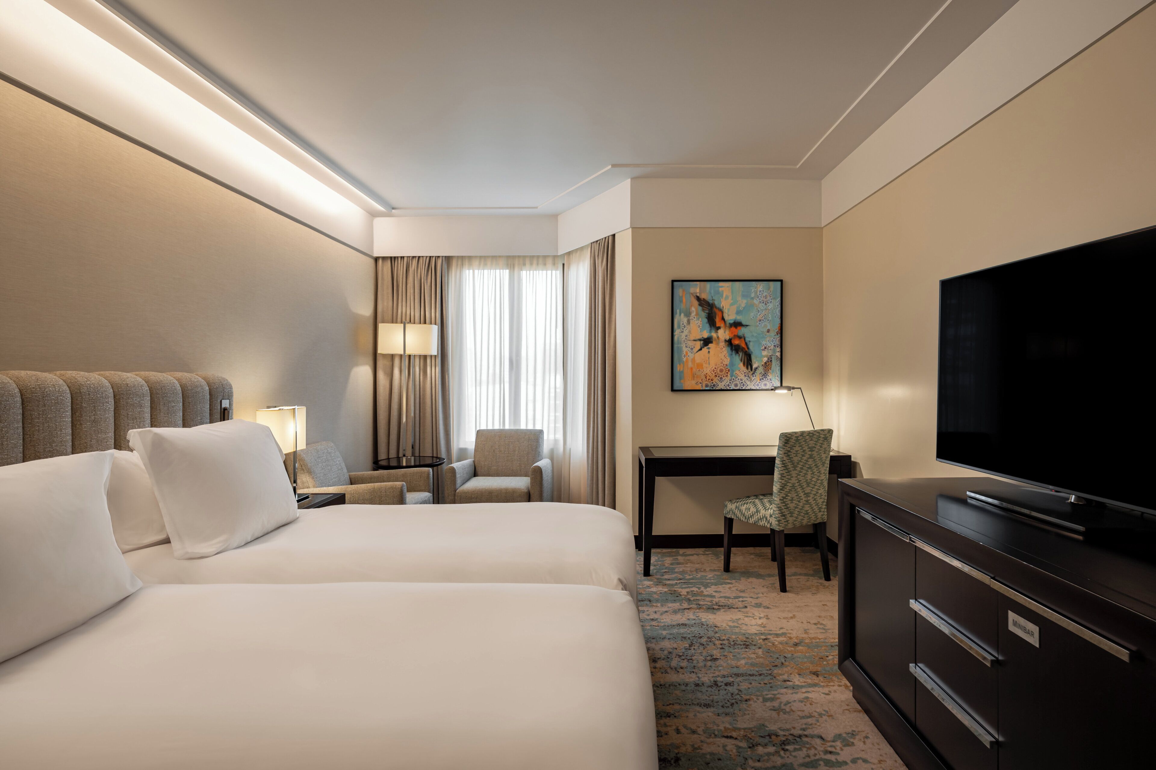 A hotel room with two beds, a desk, a chair, a television, and a painting on the wall.