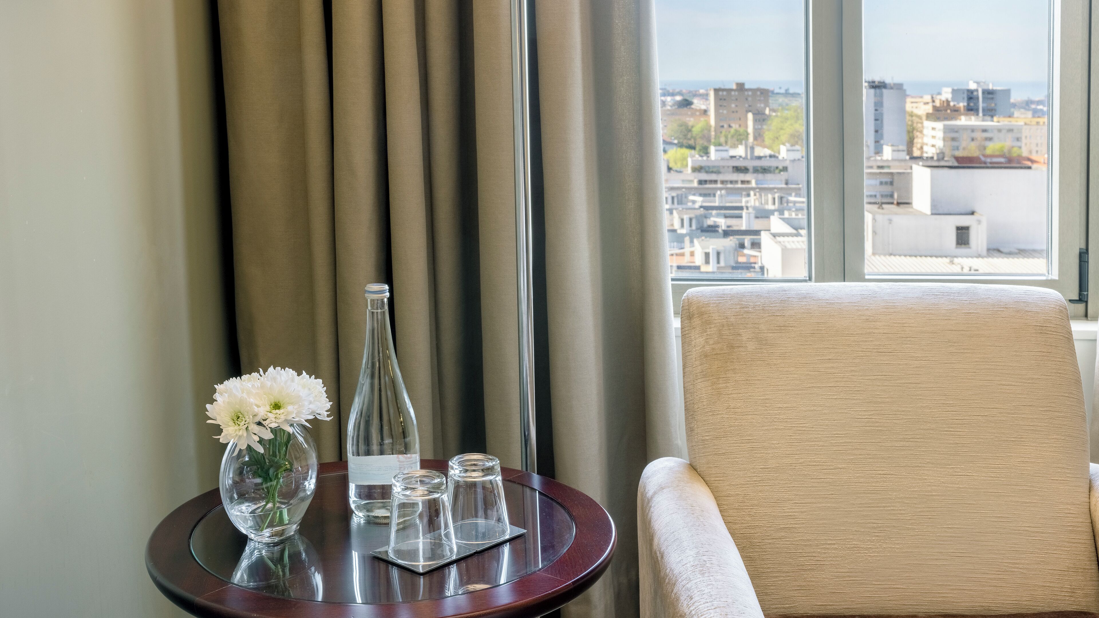 A hotel room with a view of the city, a round table with a vase of flowers, a bottle, and glasses.