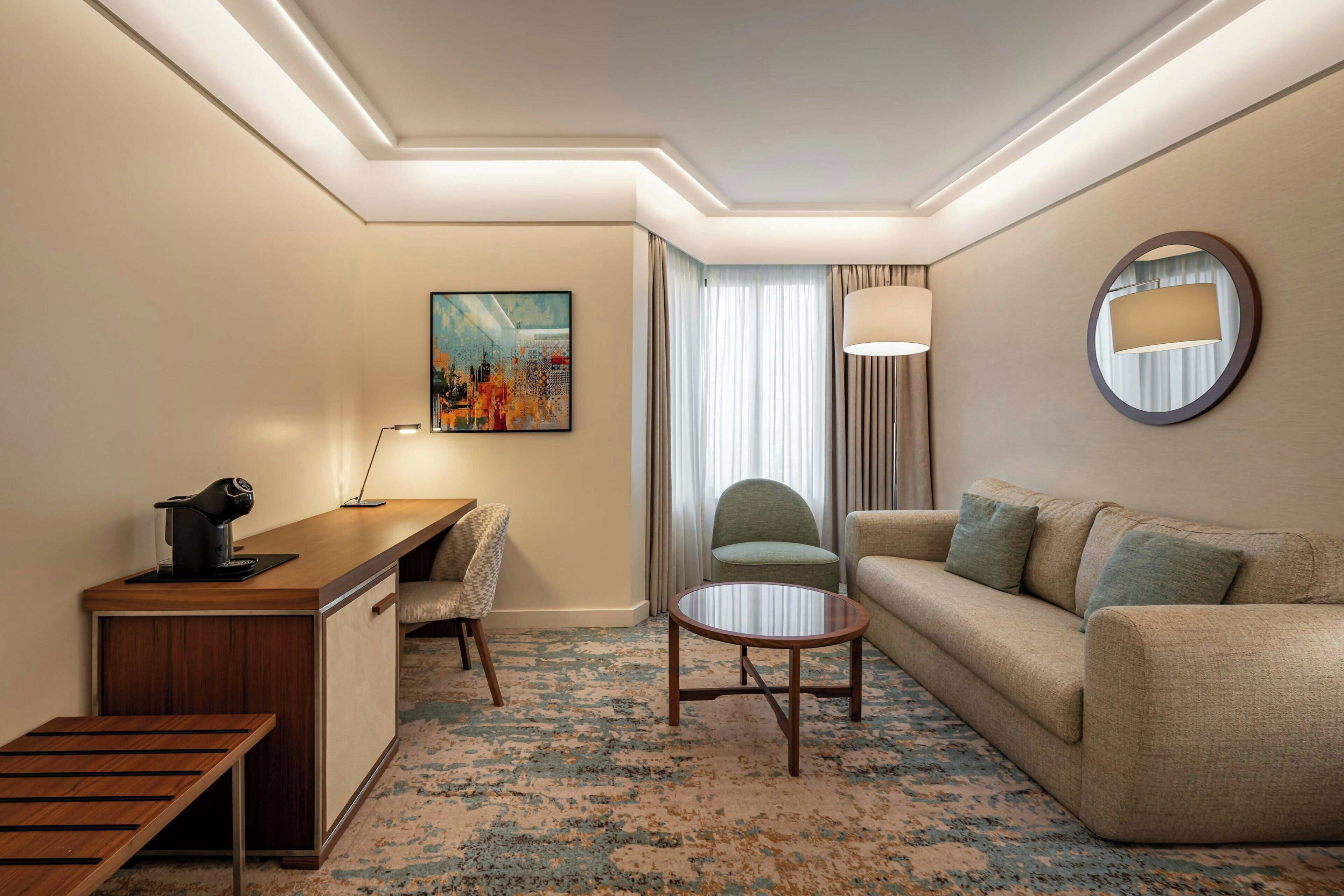 A hotel room with a desk, a sofa, a chair, a round coffee table, and a painting on the wall.
