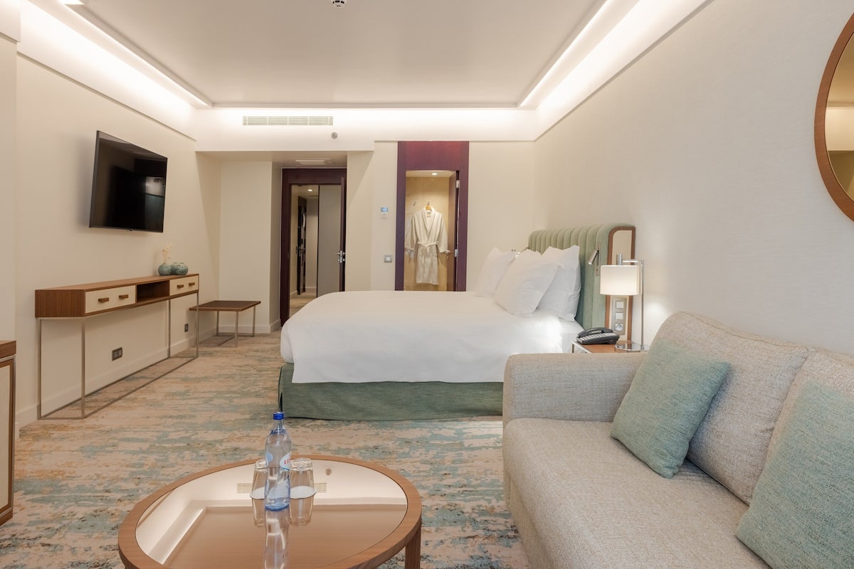 A hotel room with a bed, a sofa, a desk with a flat-screen TV, a bedside table with a lamp, and a carpeted floor.