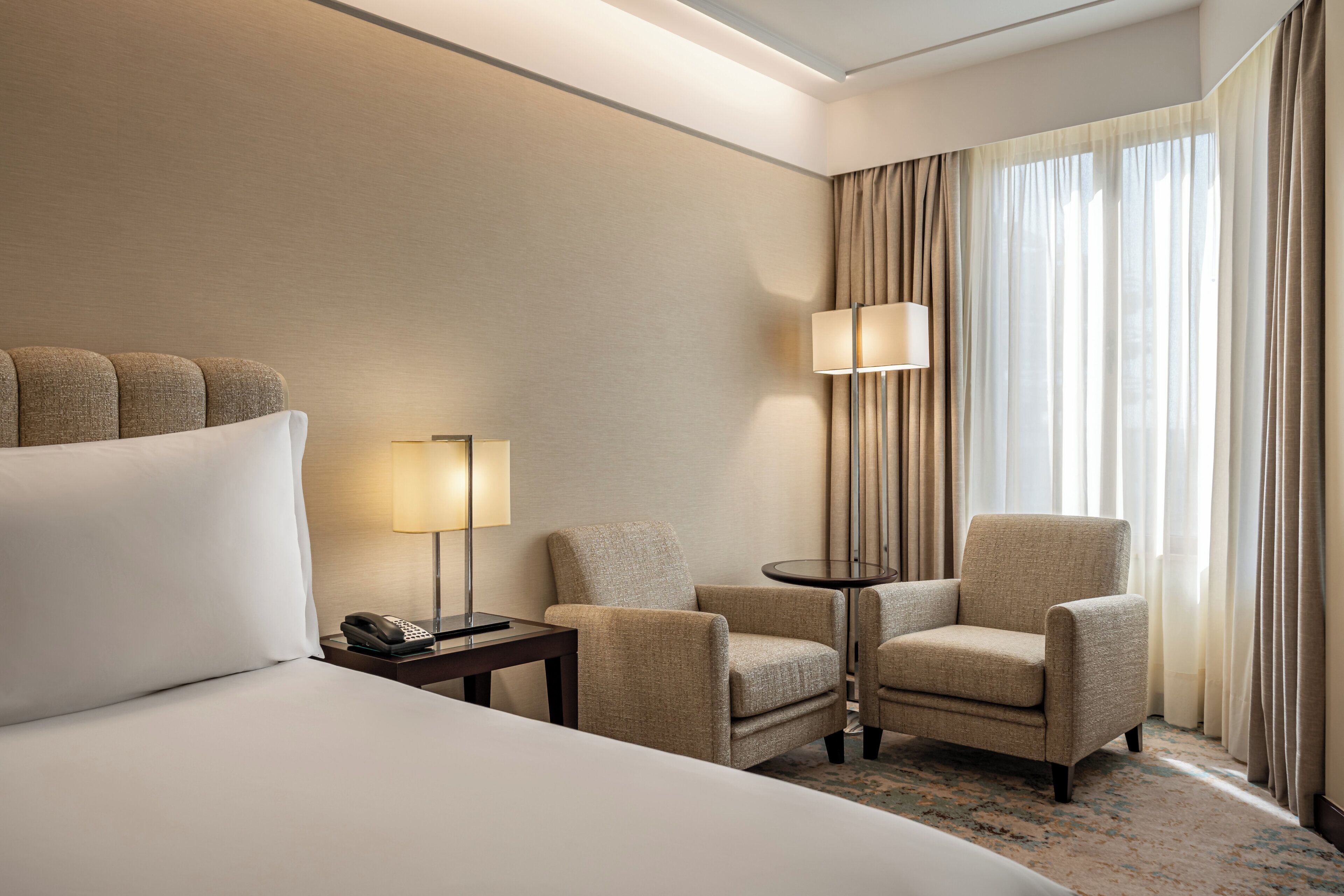 A hotel room with a large bed, two armchairs, a small table with a lamp, a telephone, and a window with curtains.